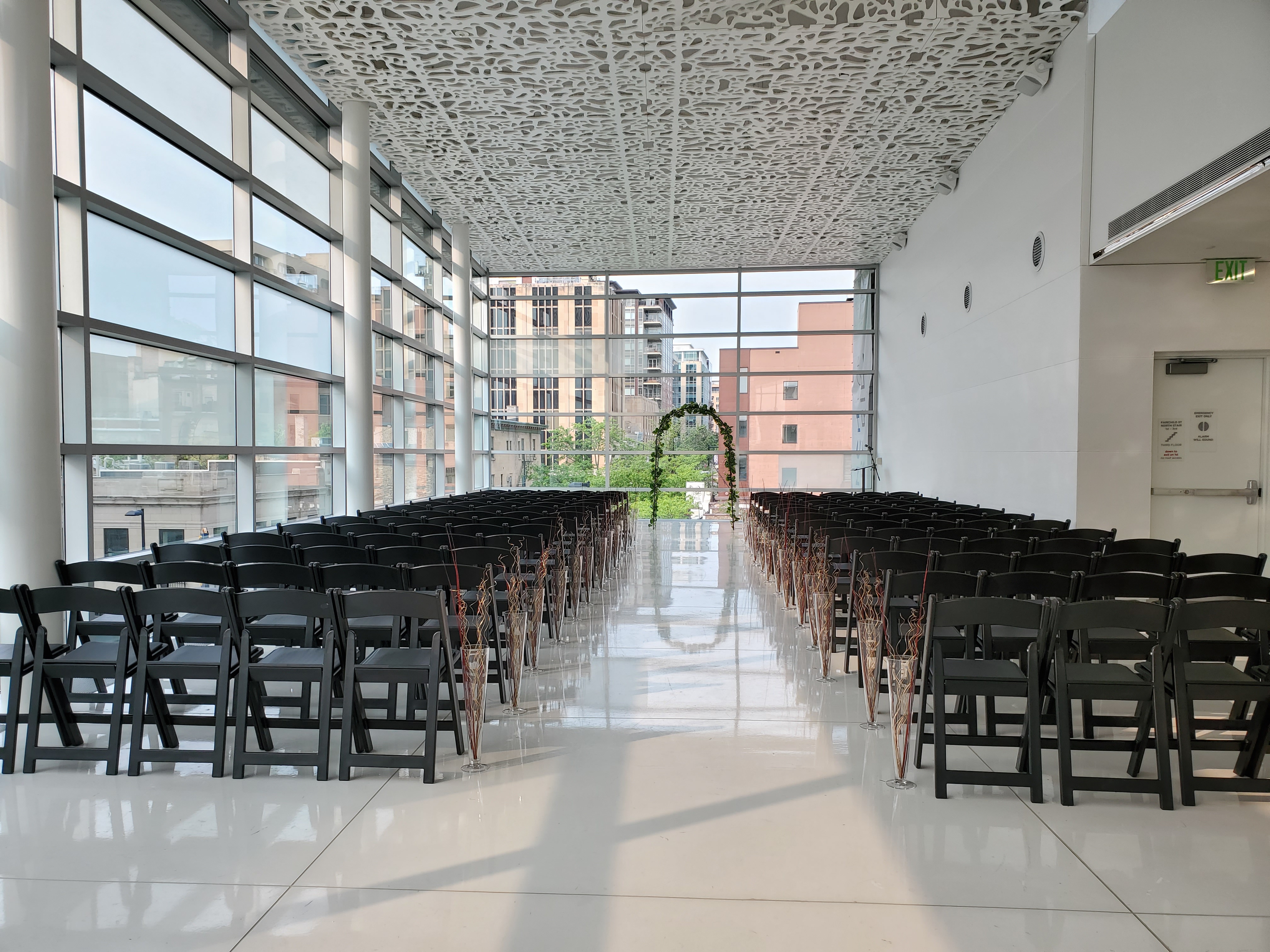 The madison room set up for a wedding ceremony with rows of black chairs and an aisle facing book arch in front of windows