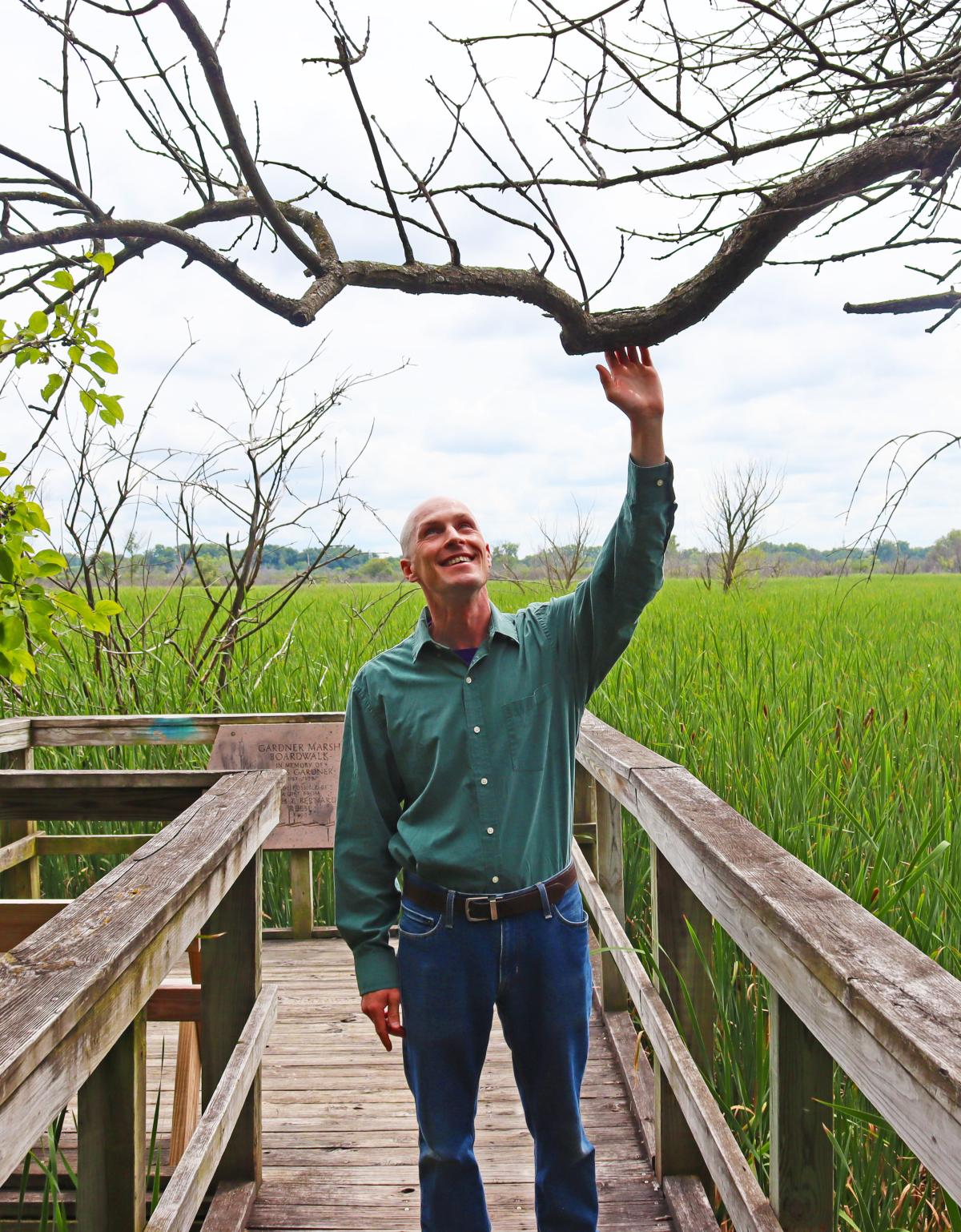 Madison Public Library Naturalist-in-Residence John C. Newman stands among the trees with a prairie in the background at the UW Arboretum