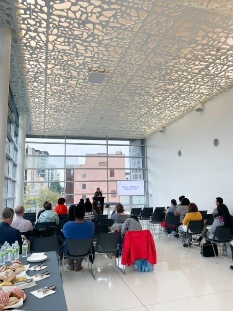 people sitting in rows watching a presenter at a podium in front of a wall of windows